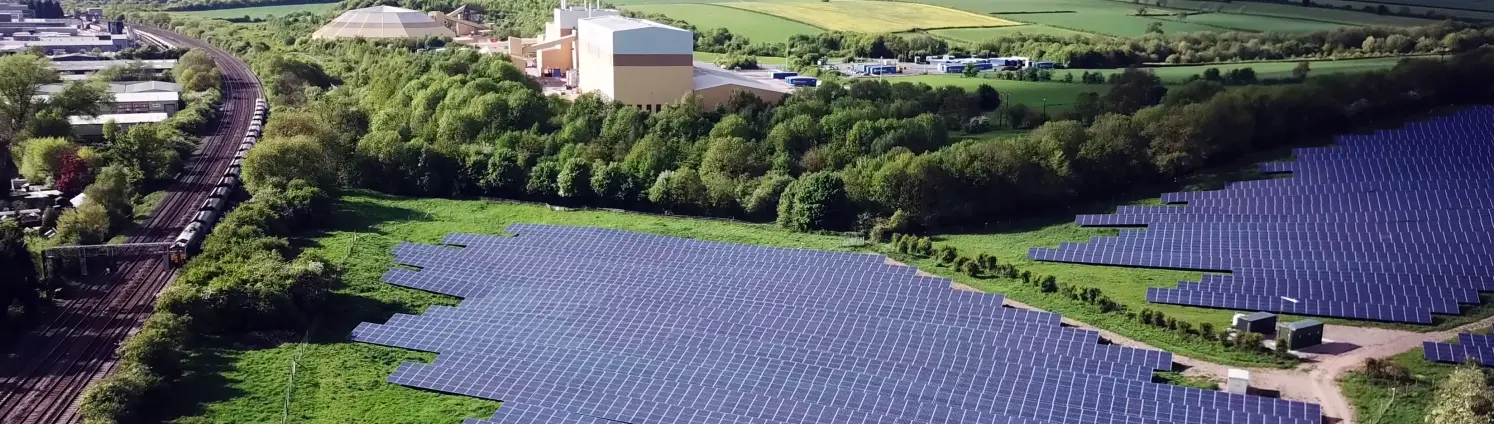 Ein ausgedehnter Solarpark neben Bahngleisen aus der Luft. Im Hintergrund Felder, Wälder, Ortschaften und Industrieanlagen.