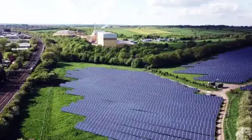Ein ausgedehnter Solarpark neben Bahngleisen aus der Luft. Im Hintergrund Felder, Wälder, Ortschaften und Industrieanlagen.