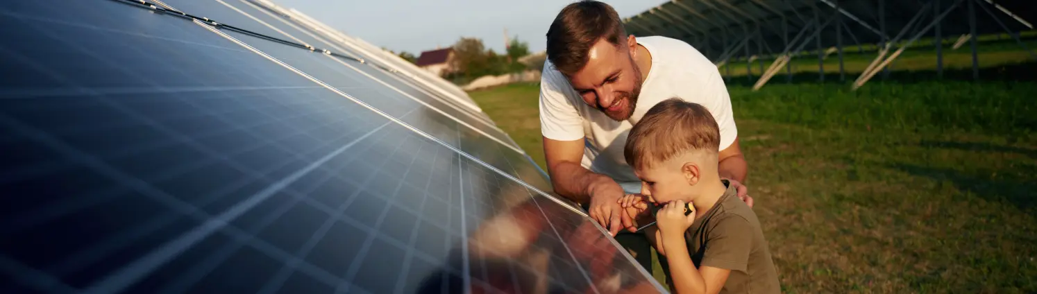 Vater und Sohn warten und reinigen gemeinsam eine lange Reihe von schräg stehenden Solarzellen auf einer Wiese.